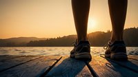 Person standing on wooden dock at sunset overlooking calm lake with mountains in background, wearing athletic shoes for outdoor recreation and fitness activities.