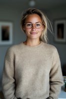 Young Caucasian woman with blonde hair wearing eyeglasses and beige sweater smiles warmly at camera. Natural lighting creates soft mood in indoor setting.