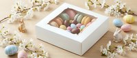 Colorful macarons in white gift box surrounded by spring cherry blossoms and pastel Easter eggs on wooden surface for holiday celebrations.