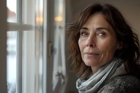 Thoughtful middle-aged Caucasian woman with brown hair gazing through window in natural light, wearing gray sweater and scarf, perfect for lifestyle or wellness advertising.