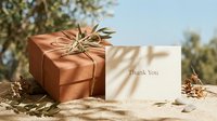 Terracotta gift box tied with twine and olive branch beside a thank you card on sandy surface with warm natural light.