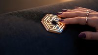Woman's hand with manicured nails and silver rings holding geometric gold pendant on dark fabric, suitable for jewelry, fashion, and luxury lifestyle marketing.