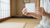 Woman in white clothing holding blank white card in minimalist interior space for mockup, branding, greeting card, or invitation design purposes.