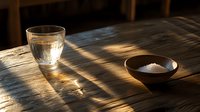 Glass of pure water and bowl with salt on wooden table in dramatic morning sunlight casting long shadows. Minimalist food composition.