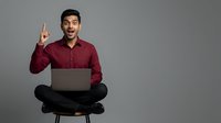 Young man with dark hair and South Asian ethnicity wearing burgundy shirt, sitting cross-legged with laptop, pointing upward with excited expression against gray background for business and technology concepts.