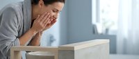 Middle-aged woman with dark hair and olive complexion experiencing fatigue or stress while working at wooden desk in bright office environment with window.