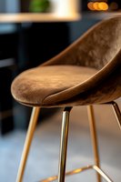 Modern brown velvet bar stool with golden metal legs captured in soft focus. Luxurious furniture detail highlighting curved seat design and premium materials.