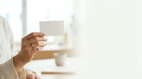 Professional holding blank white business card in hand against blurred office background. Suitable for corporate branding, business identity, and professional networking materials.