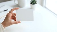 Cropped hand holding blank white business card on modern office desk with succulent plant and cup, ready for branding mockup design.