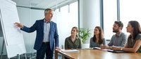 Mature male professional in business suit presenting strategy on whiteboard to diverse team of four colleagues seated at wooden conference table in modern office with large windows and natural light.