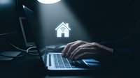 Hands typing on laptop with glowing house icon representing smart home technology, real estate search, or property management in dark workspace.