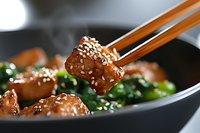 Glazed teriyaki chicken pieces with sesame seeds held by wooden chopsticks over stir-fried spinach in black pan, close-up shot showing texture and shine.