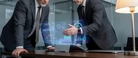 Two professional businessmen in dark suits analyzing holographic financial data and charts on modern glass table in contemporary office environment.
