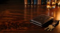Leather wallet and keys on polished wooden desk surface with whiskey glass and books in warm ambient lighting for business and lifestyle concepts.