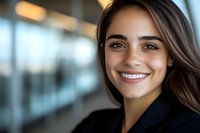 Young professional woman with radiant smile and long brown hair wearing black blazer against blurred office background, expressing confidence and success.
