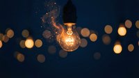 Glowing incandescent bulb with golden light particles and bokeh effect on dark blue background, suitable for innovation, creativity, and energy concepts.
