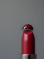 Red ladybug beetle perched on burgundy lipstick tube against gray background, minimalist beauty and nature concept for cosmetic advertising campaigns.