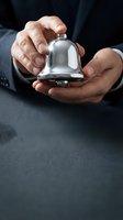 Businessman in dark suit holding shiny silver service bell over gray table, symbolizing hospitality, customer service, and luxury hotel concierge concepts.