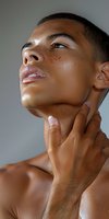 Young African American male model with freckles and glowing skin touching neck in artistic beauty portrait against neutral background.
