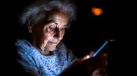 Senior Caucasian woman with gray hair using smartphone in dark room with warm lighting. Elderly person browsing mobile device at night for communication.