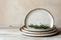 Stack of rustic ceramic speckled plates with fresh rosemary on a wooden surface, neutral beige background, minimalist kitchen decor.
