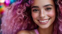 Young mixed race woman with pink curly hair and bright smile against colorful bokeh background, expressing joy and confidence in modern urban style.
