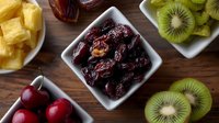 Fresh and dried fruits assortment in white ceramic bowls on wooden table including dates, cherries, pineapple and sliced kiwi, top view composition.