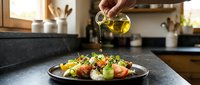 Olive oil pouring over fresh vegetable salad with tomatoes, cucumbers, and herbs on dark plate in modern kitchen setting.