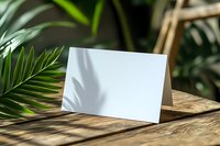 Blank white folded card on wooden table with tropical palm leaves in background, creating natural ambiance for invitation or greeting card design.