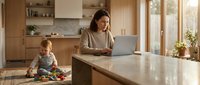 Young mother working on laptop in modern kitchen while toddler plays nearby. Remote work and parenting balance concept for busy families.