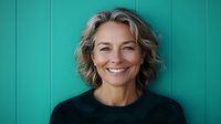 Confident mature woman with blonde wavy hair wearing black sweater smiling genuinely against turquoise wall background, representing wellness and positive aging concept.