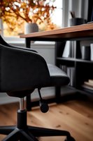 Modern home office workspace with black ergonomic chair and wooden desk against window, autumn trees visible outside creating warm atmospheric setting.
