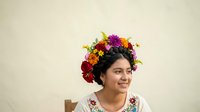Young woman with dark hair and medium skin tone wearing a vibrant floral crown of red, purple, and yellow flowers. She wears an embroidered white top and smiles while looking to the side against a neutral beige background. Suitable for lifestyle, beauty, and cultural content.