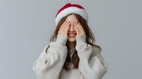 Young woman in Santa hat covering eyes with hands while smiling broadly, wearing cozy white sweater against neutral gray background.