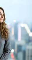 Professional woman with long brown hair wearing gray blazer against blurred cityscape background with bokeh lights. Corporate portrait for business and leadership concepts.