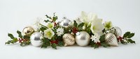 Elegant Christmas centerpiece with white amaryllis flowers, silver ornaments, holly berries and greenery arranged on white surface for holiday decor.