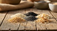 Assorted seeds and grains arranged in small piles on a rustic wooden surface, including sesame, poppy, and oat flakes, with bowls in background.