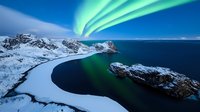 Spectacular aurora borealis over snow-covered coastal landscape with dramatic green lights dancing across night sky above dark waters and rugged mountain peaks.
