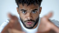 Close up portrait of young African American man with blue eyes and beard expressing surprise and disbelief, reaching hands toward camera with dramatic lighting.