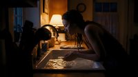 Young woman with dark hair and medium skin tone, wearing casual sleeveless top, leaning over desk in dimly lit room with warm lamp light, focused on work or study at night, suitable for education and productivity concepts.