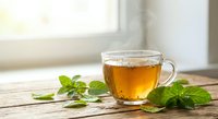 Clear glass cup of herbal tea with fresh mint leaves on wooden table by window. Natural lighting creates warm atmosphere for relaxation and wellness.