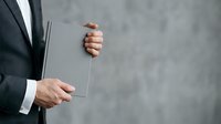 Businessman in dark suit holding blank transparent glass panel against neutral gray background, showcasing product display or corporate presentation concept.