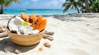 Wooden bowl with coconut, papaya slices and tropical fruits on sandy beach with turquoise ocean, palm trees and blue sky, perfect for summer travel and vacation themes.