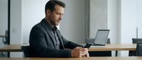 Middle-aged Caucasian male professional in dark blazer working at wooden desk with laptop and smartphone in modern office environment.