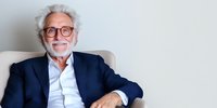 Senior Caucasian businessman with gray hair, glasses and beard wearing navy suit sits relaxed in armchair against white wall, expressing confidence and success.