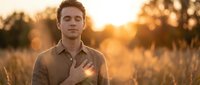 Young man with light brown hair and fair complexion wearing a tan button-up shirt standing in golden wheat field during sunset, hand on chest, contemplative expression, warm natural lighting.