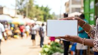 Woman with dark skin tone wearing a teal top and gold bracelets holds a blank white card at an outdoor market or community gathering, ideal for promotional messaging and advertising campaigns.