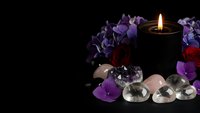 Dark candle burning with purple flowers, crystals and gemstones arranged for spiritual meditation, wellness rituals and relaxation on black background.