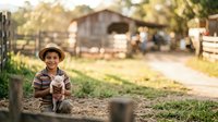 Young boy wearing straw hat and plaid shirt holding piglet on rural farm with barn and fencing in background during daytime.
