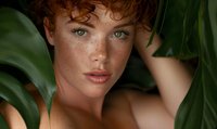Young woman with red curly hair and freckles surrounded by green tropical leaves, creating natural beauty portrait for skincare advertising.